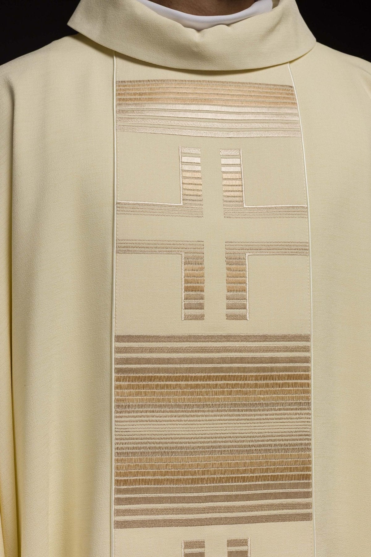 Casula creme em close-up frontal, destacando a cruz e as faixas bordadas