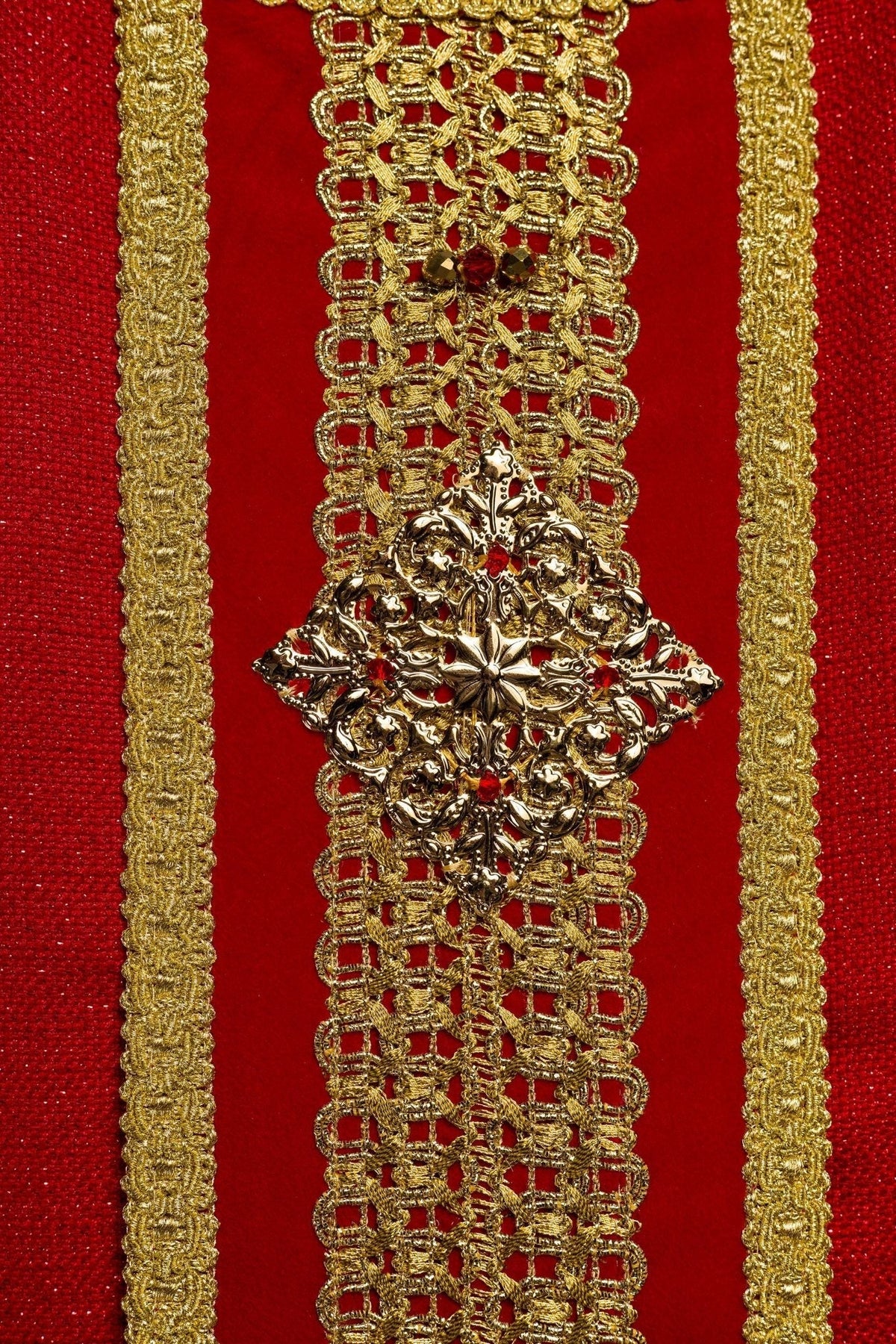 Casula vermelha em close-up, com faixa central dourada, pedras naturais e ornamento metálico