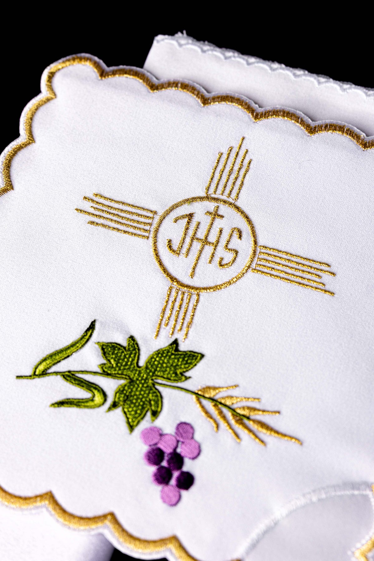 Alfaias litúrgicas brancas em close-up, com monograma IHS dourado e bordado de uvas