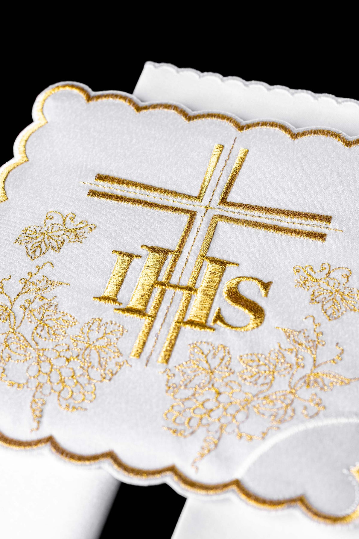 Alfaia litúrgica branca em close-up, com cruz dourada, monograma IHS e uvas bordadas