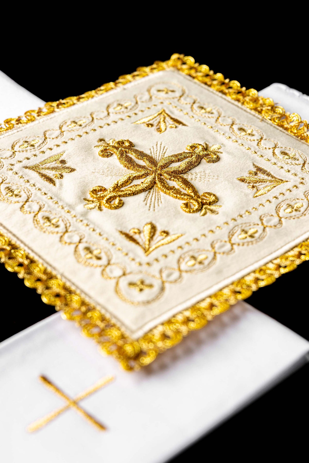Conjunto de alfaias litúrgicas em veludo creme, close-up da cruz bordada e acabamento dourado