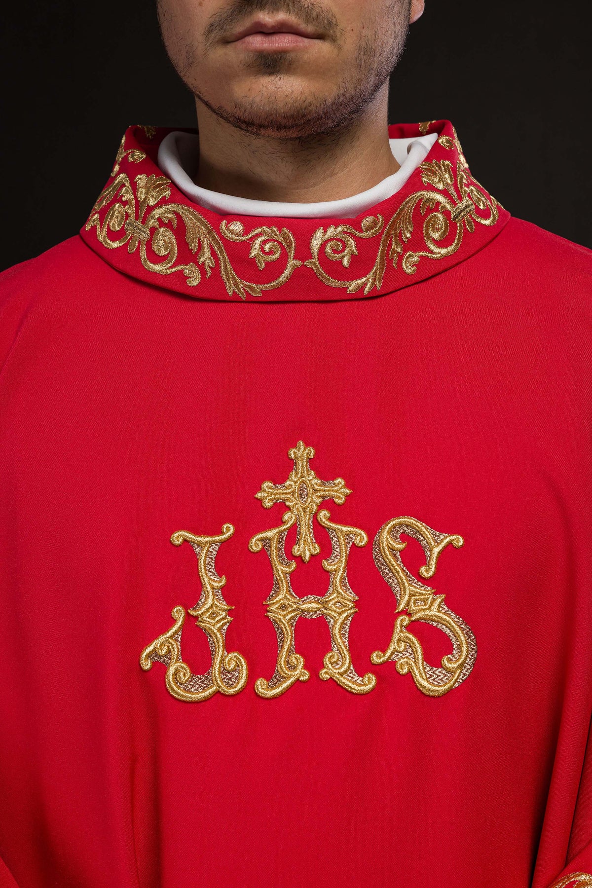 Casula vermelha em close-up frontal, destacando o bordado IHS dourado e a gola ornamentada