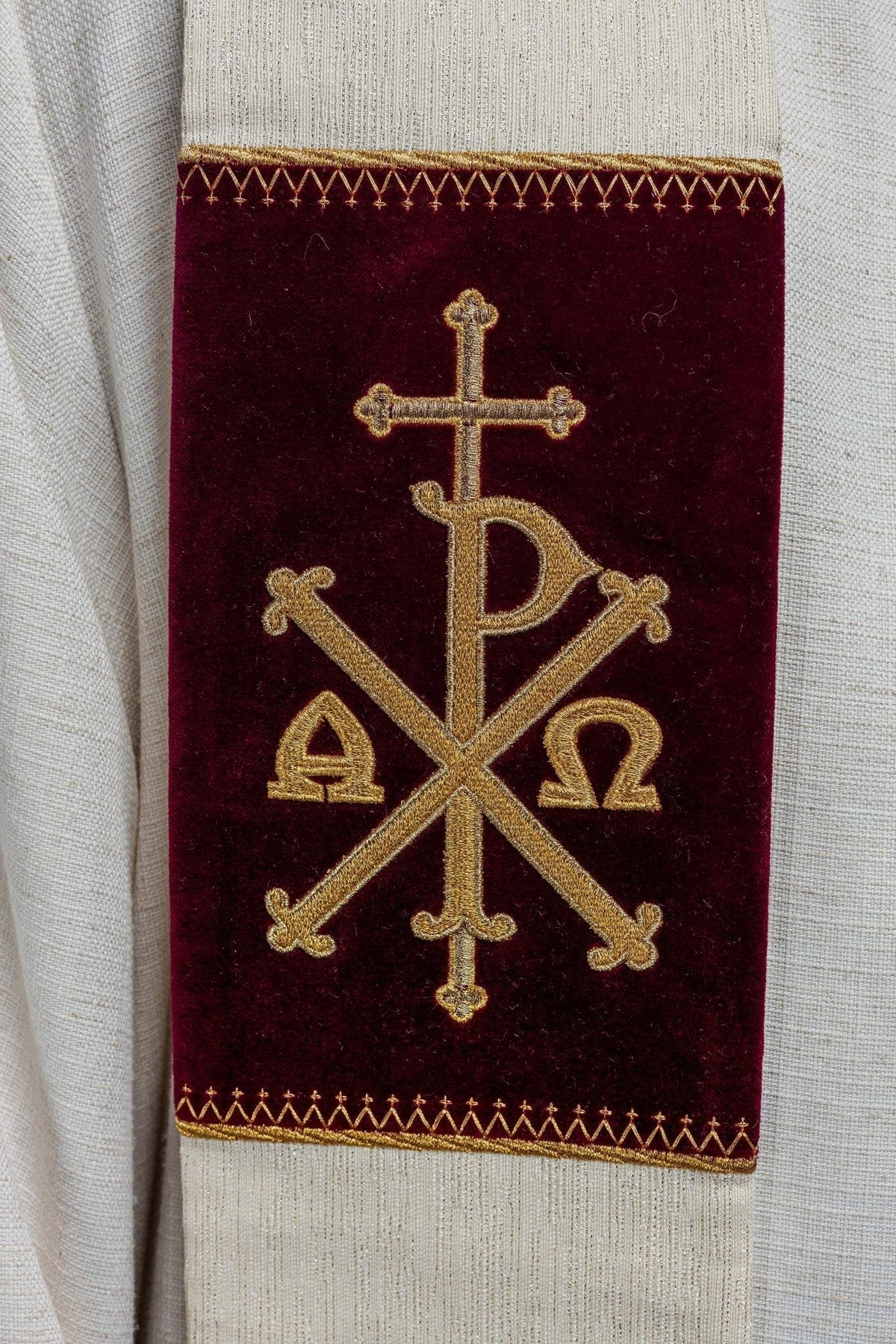 Estola ecru com close-up do bordado dourado PAX e letras Alfa e Ômega sobre faixa bordô