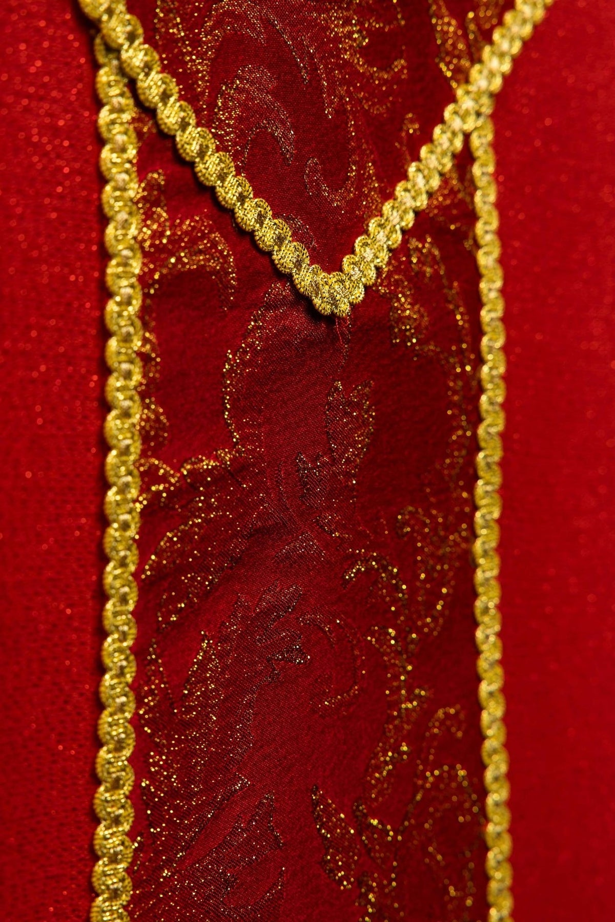 Casula vermelha em close-up, mostrando cruz dourada sobre tecido jacquard com arabescos