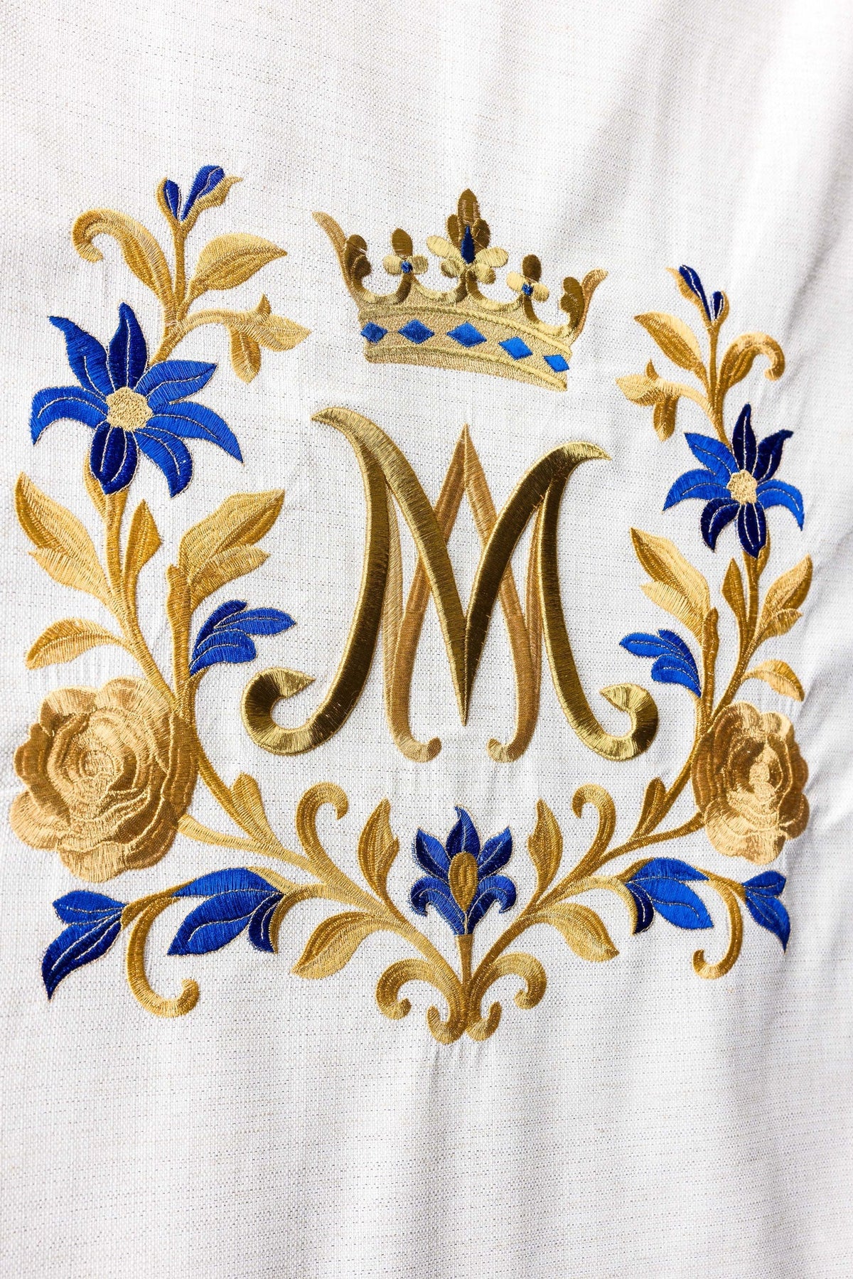 Pano para ambão creme em close-up, bordado mariano dourado e azul com monograma M e flores