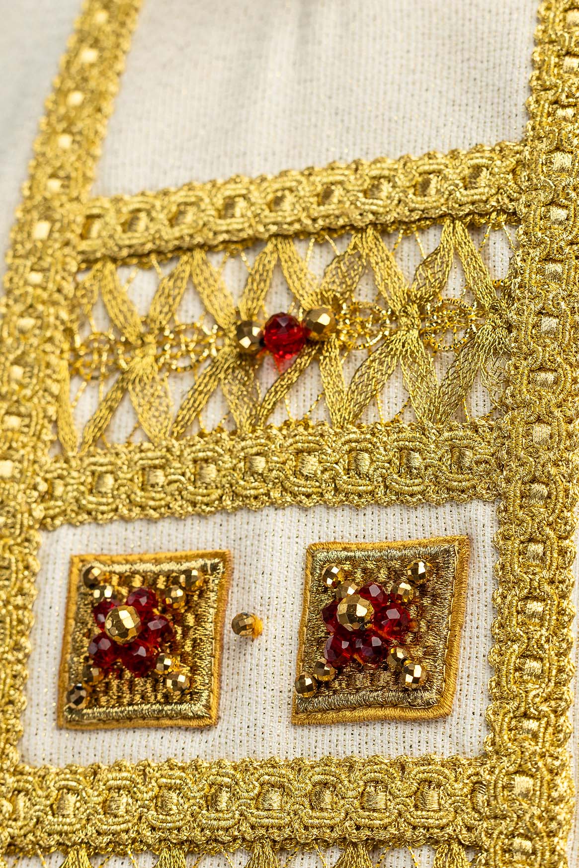 Casula artesanal em close-up da faixa frontal, com renda dourada vazada e pedra vermelha