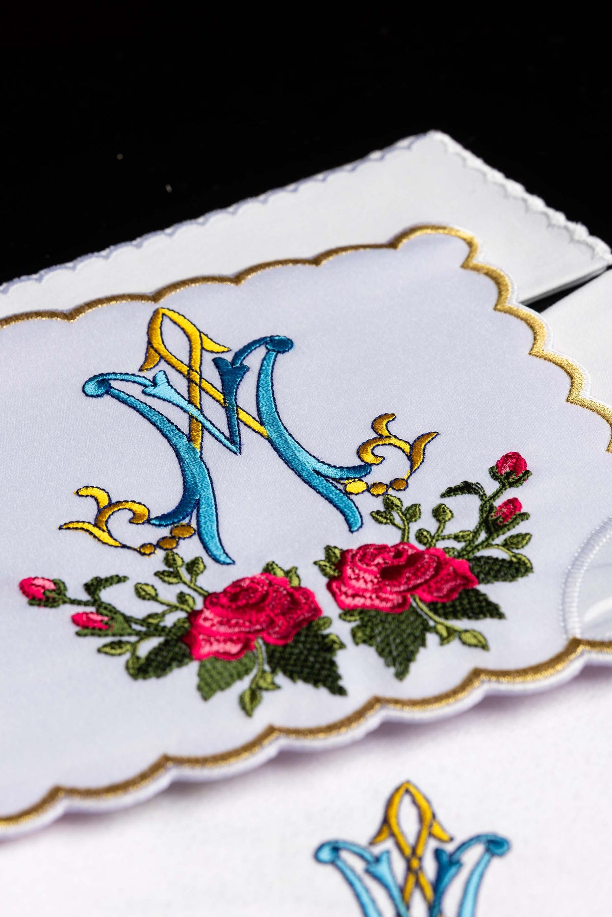 Alfaias litúrgicas brancas em close-up, com monograma mariano azul e dourado e rosas vermelhas