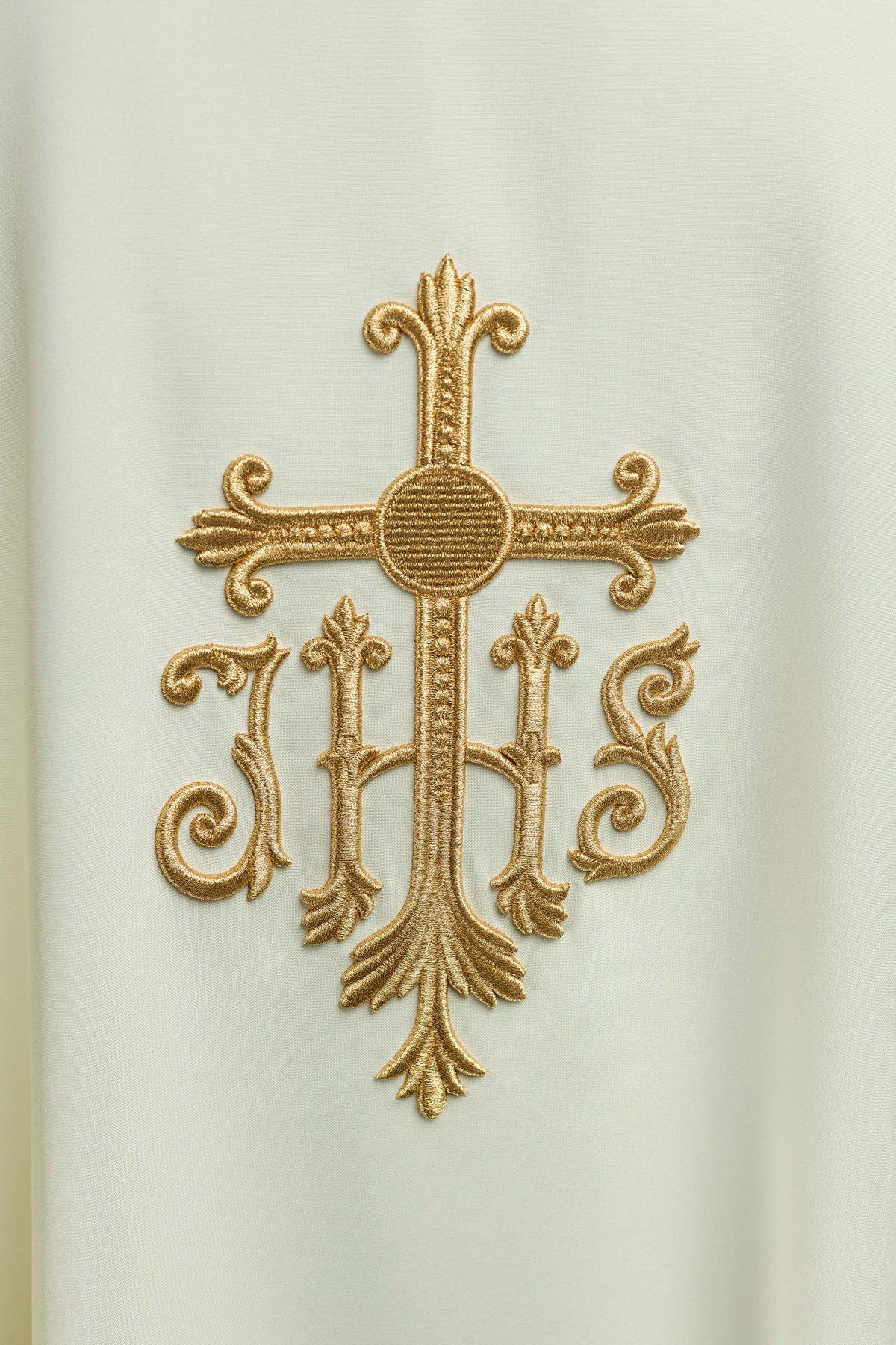 Casula creme com bordado IHS em close-up, mostrando o monograma dourado