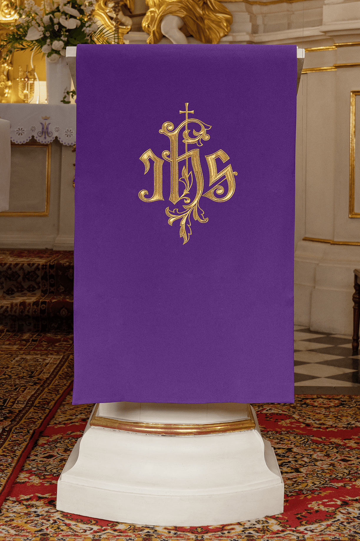 Pano de ambão roxo com bordado dourado do monograma IHS coroado por uma cruz e ornamentos vegetais, pendurado em um ambão na igreja.