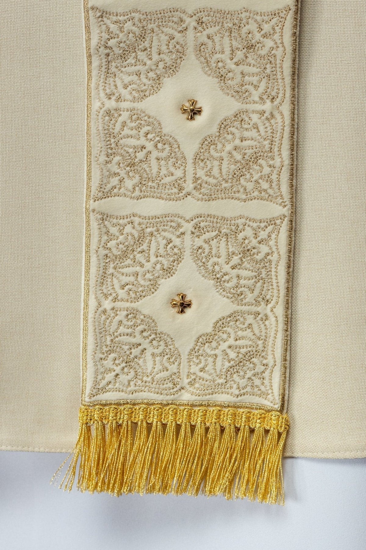 Casula creme, close da faixa bordada com motivos ornamentais, cruzes e franjas douradas